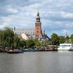 Blick auf die Leeraner Innenstadt mit dem historischen Museumshafen, Segelbooten und dem markanten Rathausturm am Wasser bei sonnigem Wetter.
