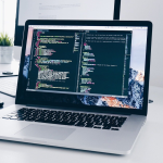 Das Bild zeigt einen geöffneten Laptop auf einem Schreibisch. Auf dem Bildschirm ist ein Programmierquellcode zu sehen.