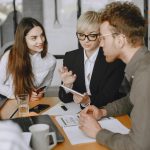 Das Bild zeigt zwei junge Frauen und einen Mann an einem Büroschreibtisch wärend eines Arbeitsmeetings.