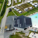 Das Bild zeigt aus der Vogelperspektive eine PV-Anlage auf dem Dach des Gebädes zum Meerwasserfreibades in Dornumersiel. Zusehen sind auch Wohnmobile, ein Spielplatz und das Meerwasserfreibad.